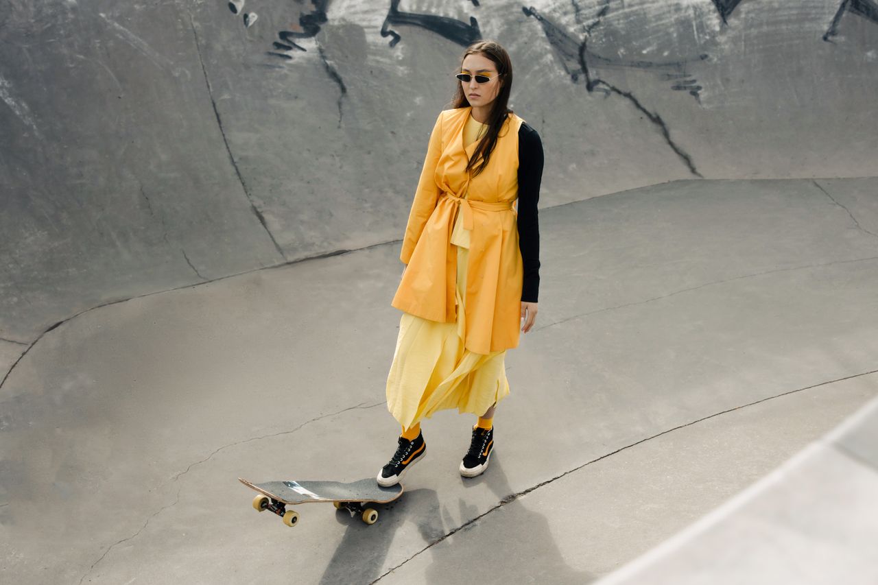 Woman in yellow dress with skateboard posing stylishly at a skate park.