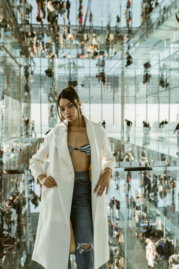 Woman in designer coat poses amidst modern mirrored architecture in New York City.